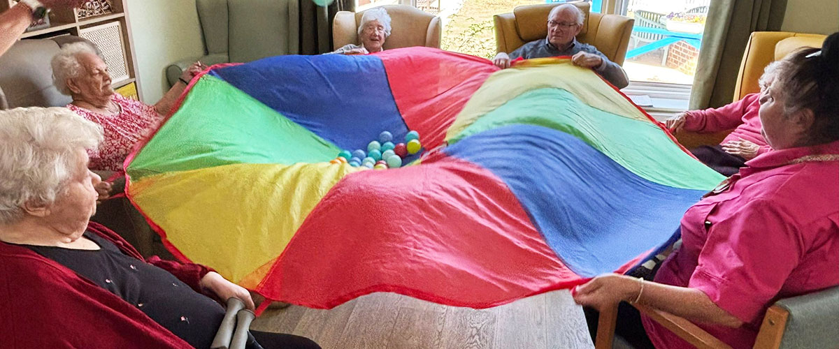 Group of Sonya Lodge residents playing a game together in their lounge, with colourful balls at the centre of a large vibrant parachute.