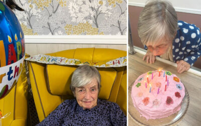 Resident Monica celebrating her birthday at Sonya Lodge, surrounded by balloons and banners, smiling as she blows out the candles on her pink floral birthday cake.